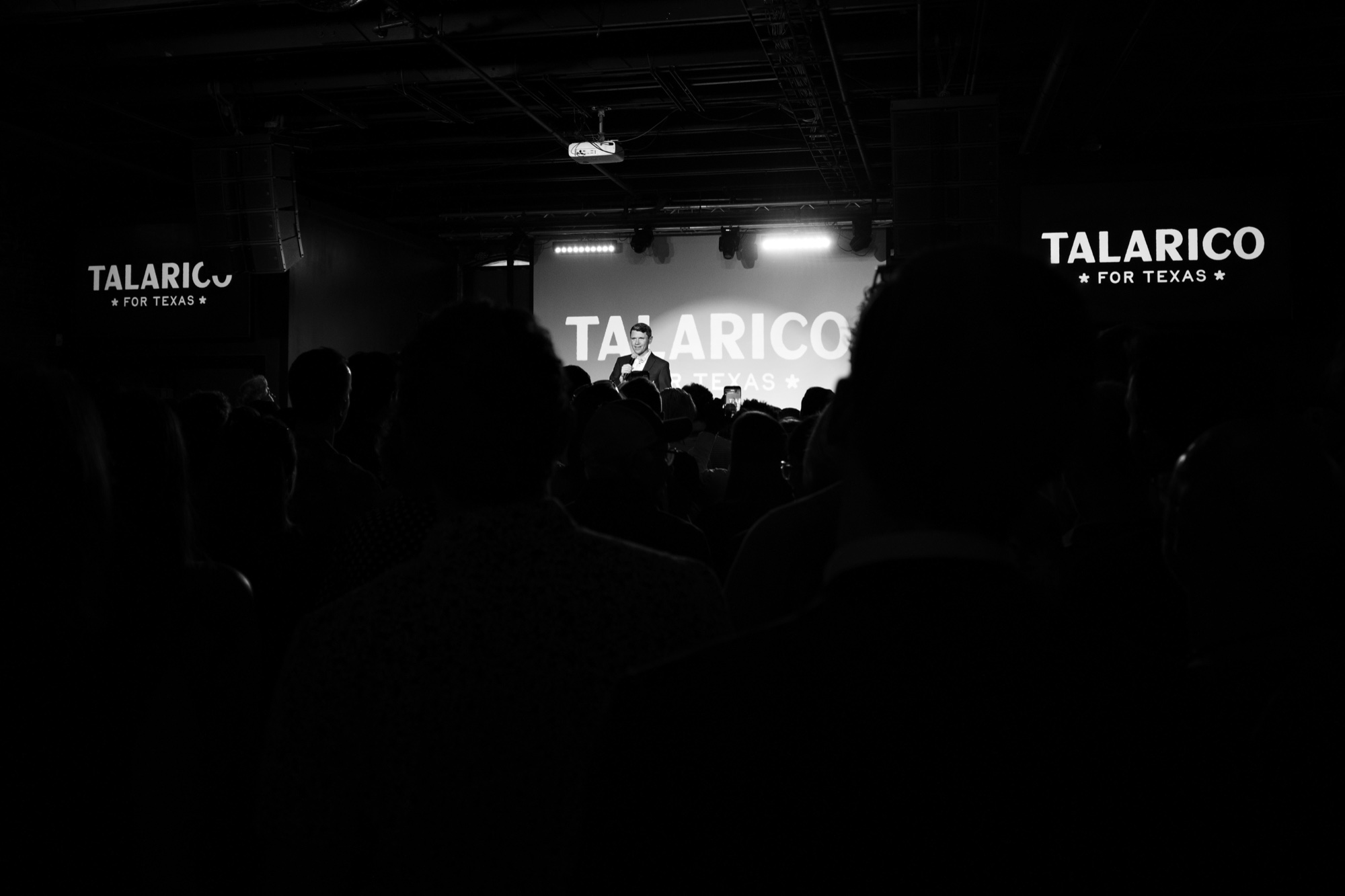 James Talarico on stage at his last Austin fundraiser before the election, October 23, 2026, seen from the back of a packed, dark room under TALARICO FOR TEXAS signs